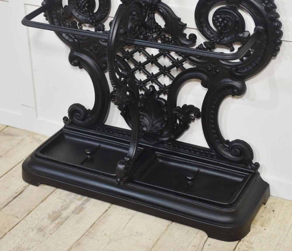 An ornate black cast iron hall stand featuring intricate scrollwork, coat hooks, an umbrella holder, and a fitted drip tray. View VI