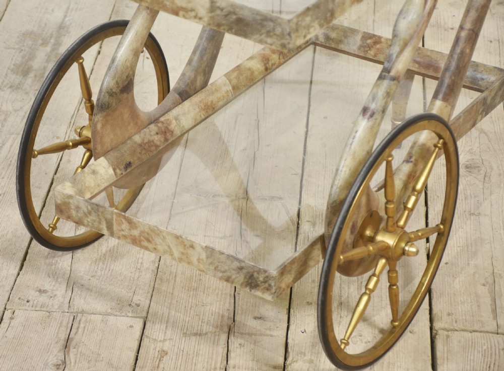 An Italian mid-century drinks trolley crafted with a lacquered goatskin frame, two glass shelves, brass-spoked wheels, and curved handles. View X