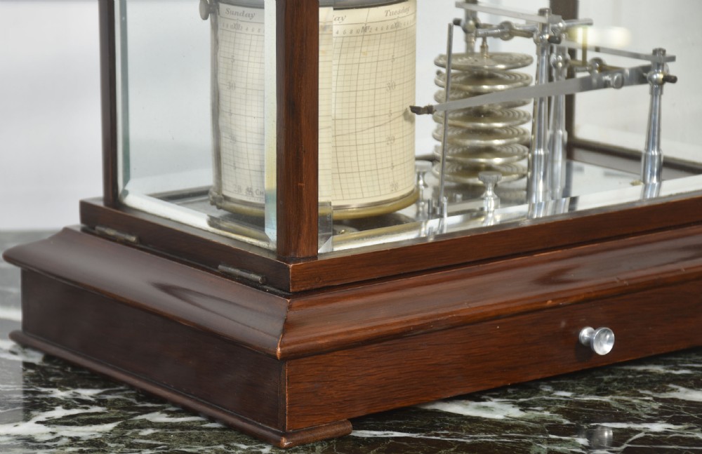 An antique mahogany cased barograph featuring bevelled glass panels enclosing a brass recording mechanism and paper chart drum above a base drawer. View II