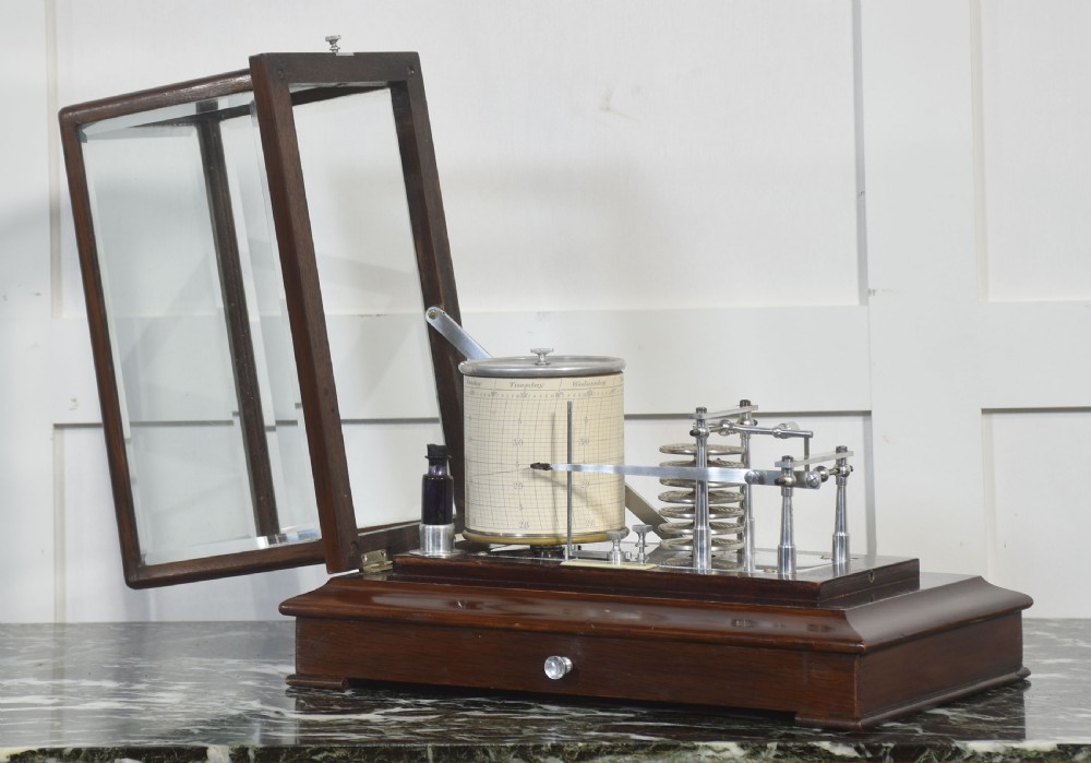 An antique mahogany cased barograph featuring bevelled glass panels enclosing a brass recording mechanism and paper chart drum above a base drawer. View IV