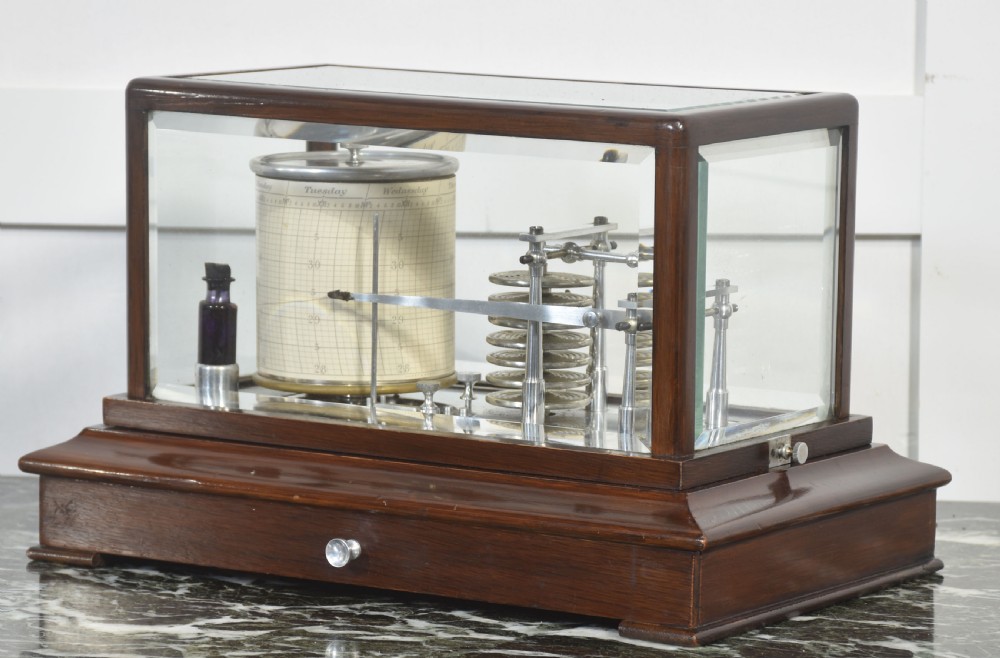 An antique mahogany cased barograph featuring bevelled glass panels enclosing a brass recording mechanism and paper chart drum above a base drawer. View VI