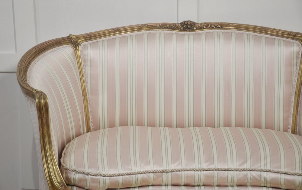 A French Louis XV style giltwood two-seater settee featuring carved floral decorations, curved arms, cabriole legs, and pink and cream striped fabric cushions. View V