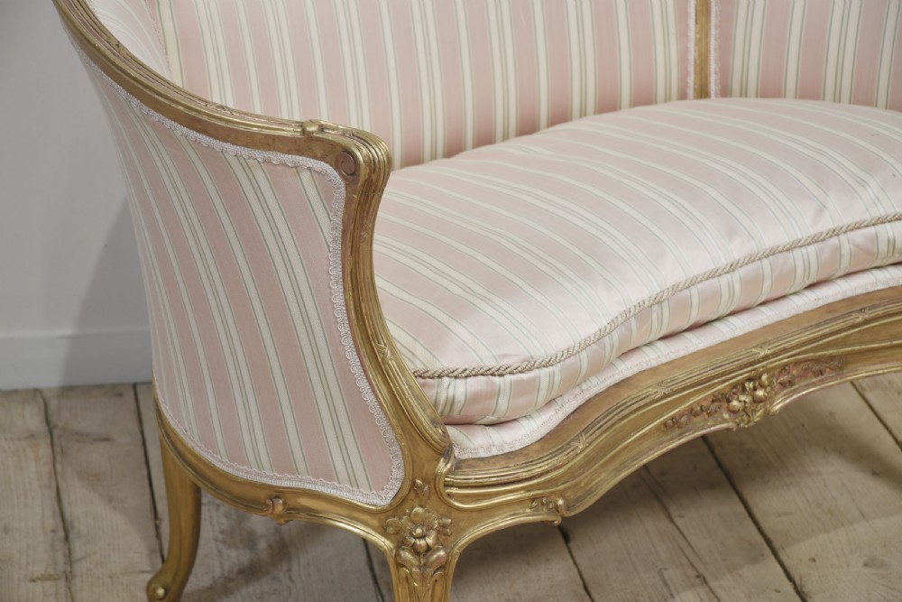 A French Louis XV style giltwood two-seater settee featuring carved floral decorations, curved arms, cabriole legs, and pink and cream striped fabric cushions. View X