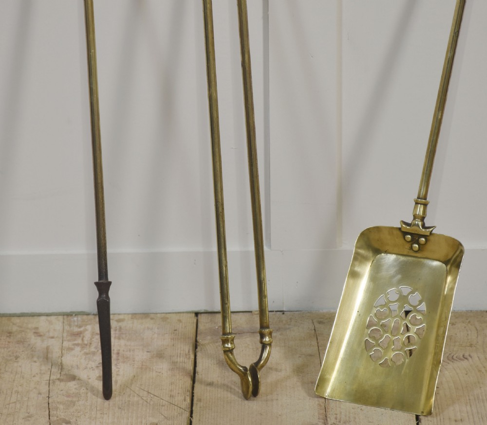 A set of three antique brass fireside tools including a poker, tongs, and a pierced shovel displayed upright. View II