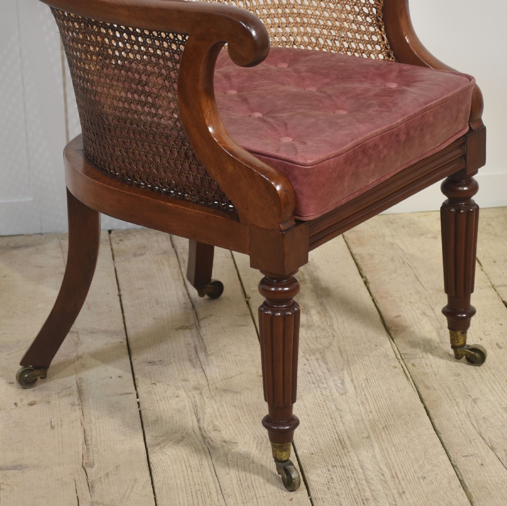 A Regency mahogany bergere armchair featuring a woven cane backrest and sides, reeded front legs on brass castors, and a buttoned red leather cushion. View II