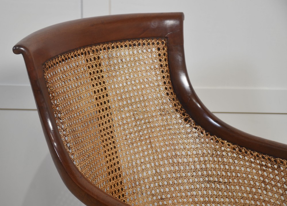 A Regency mahogany bergere armchair featuring a woven cane backrest and sides, reeded front legs on brass castors, and a buttoned red leather cushion. View III