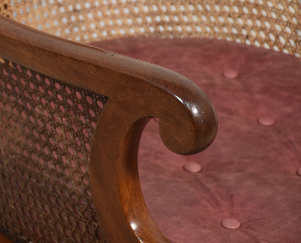 A Regency mahogany bergere armchair featuring a woven cane backrest and sides, reeded front legs on brass castors, and a buttoned red leather cushion. View V