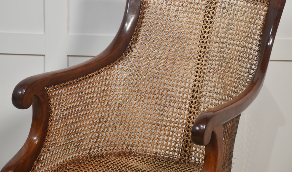 A Regency mahogany bergere armchair featuring a woven cane backrest and sides, reeded front legs on brass castors, and a buttoned red leather cushion. View VII
