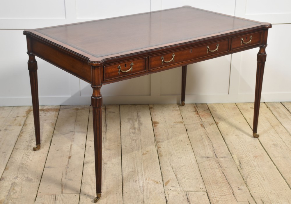An antique mahogany writing table featuring three frieze drawers with brass handles, a leather inset top, and slim reeded legs with brass castors.