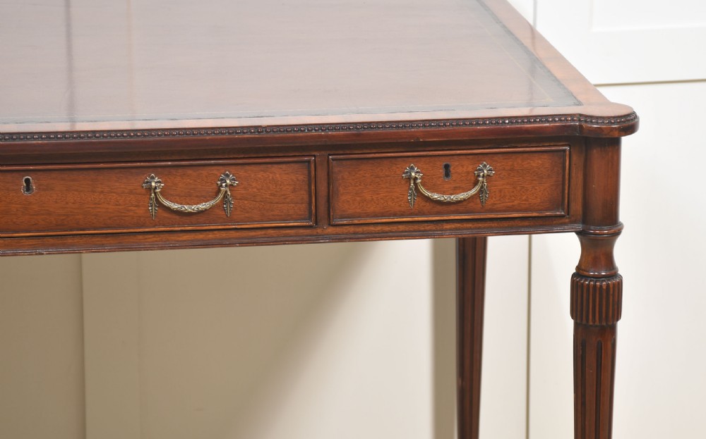 An antique mahogany writing table featuring three frieze drawers with brass handles, a leather inset top, and slim reeded legs with brass castors. View IV