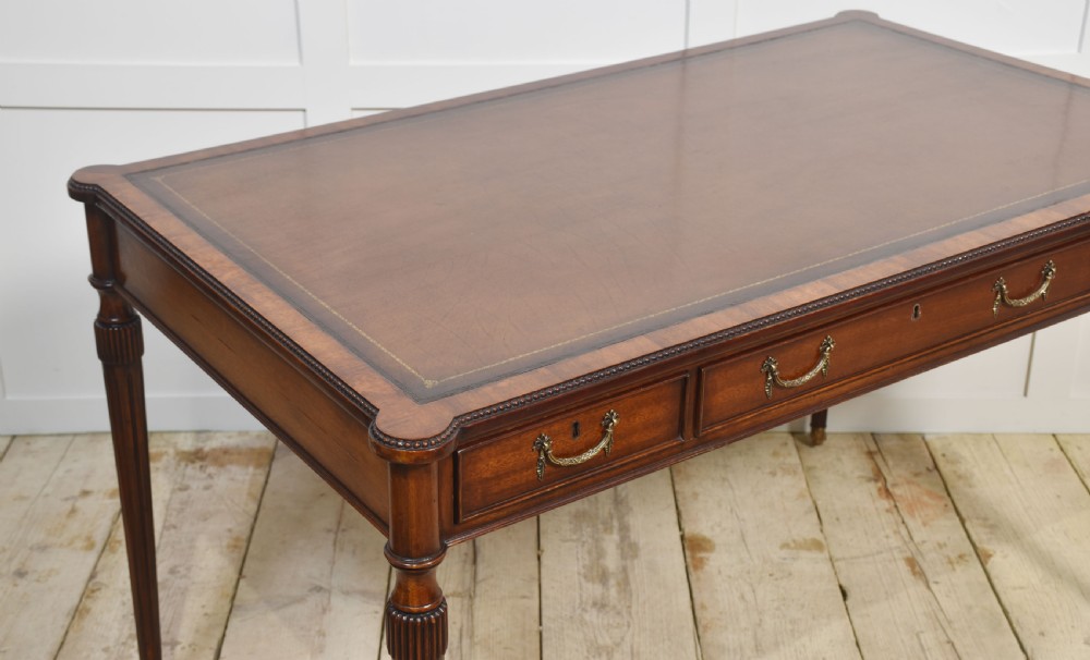 An antique mahogany writing table featuring three frieze drawers with brass handles, a leather inset top, and slim reeded legs with brass castors. View VI