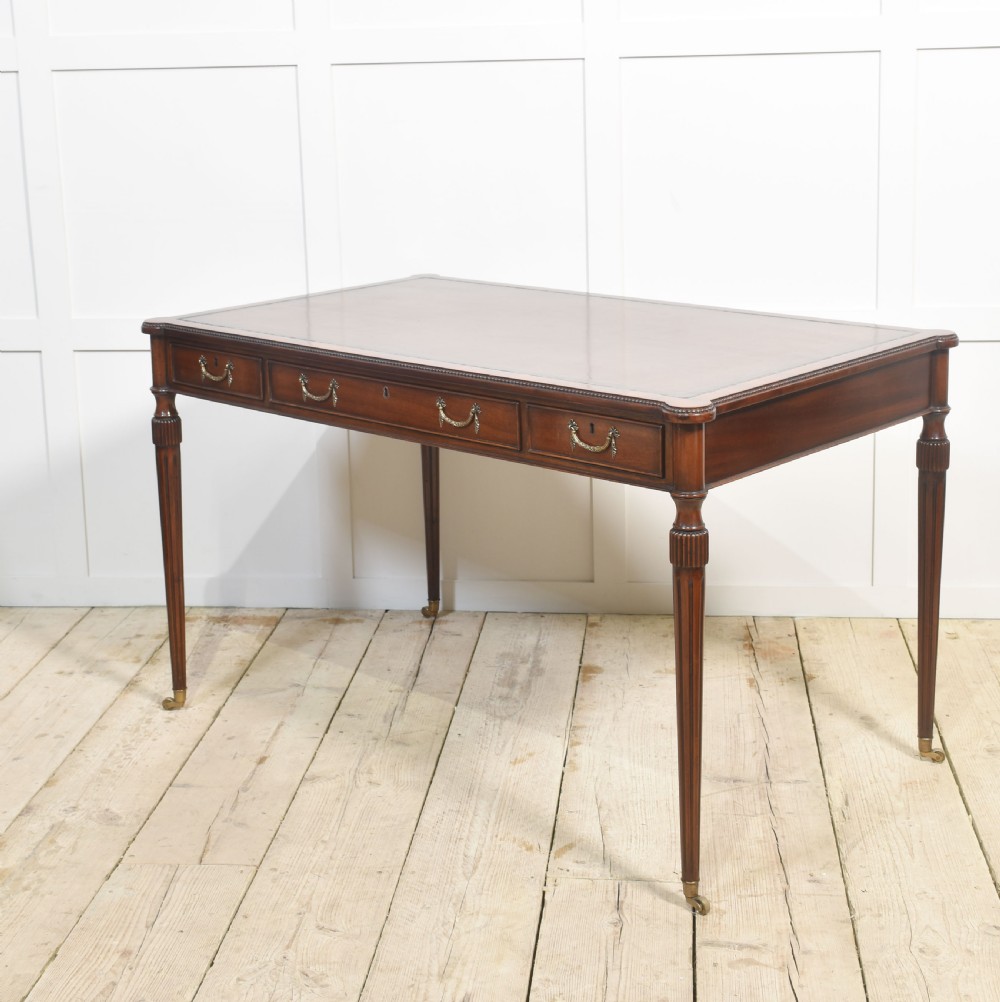 An antique mahogany writing table featuring three frieze drawers with brass handles, a leather inset top, and slim reeded legs with brass castors. View IX