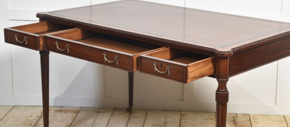 An antique mahogany writing table featuring three frieze drawers with brass handles, a leather inset top, and slim reeded legs with brass castors. View XI