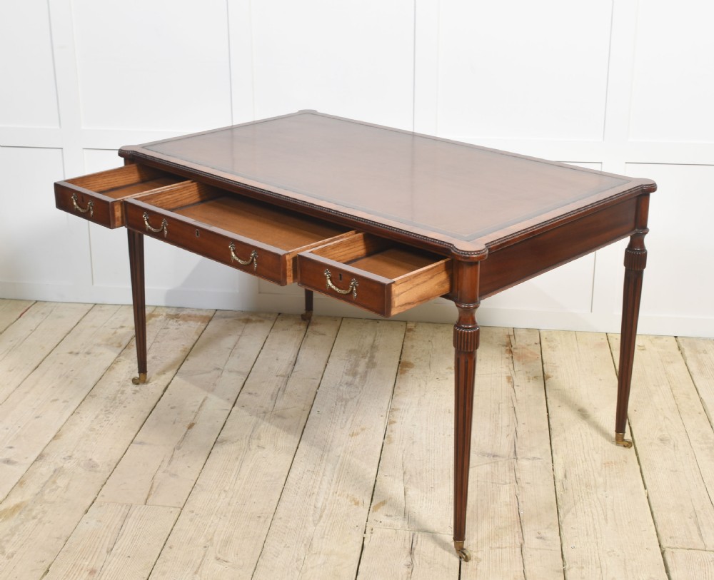 An antique mahogany writing table featuring three frieze drawers with brass handles, a leather inset top, and slim reeded legs with brass castors. View XII