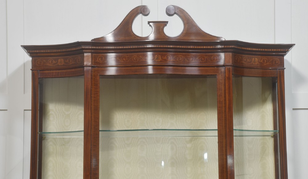 An Edwardian inlaid mahogany display cabinet featuring a swan-neck pediment, glass shelves, inlaid floral marquetry doors, and slender tapered legs. View II
