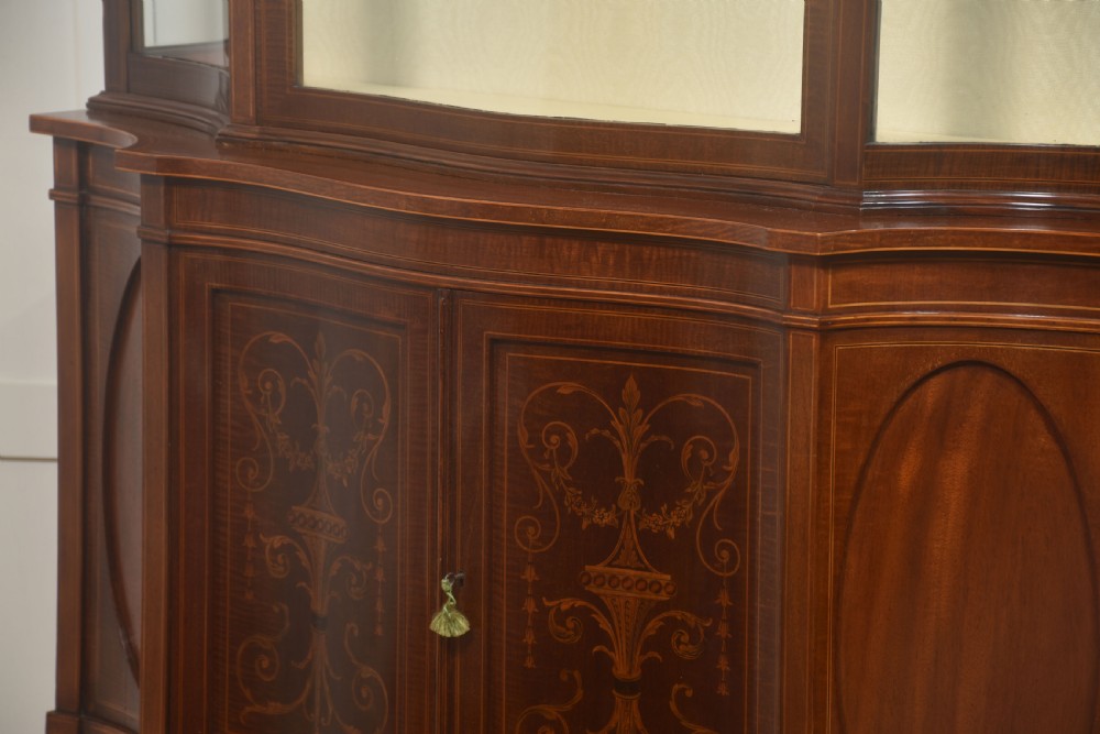 An Edwardian inlaid mahogany display cabinet featuring a swan-neck pediment, glass shelves, inlaid floral marquetry doors, and slender tapered legs. View IX