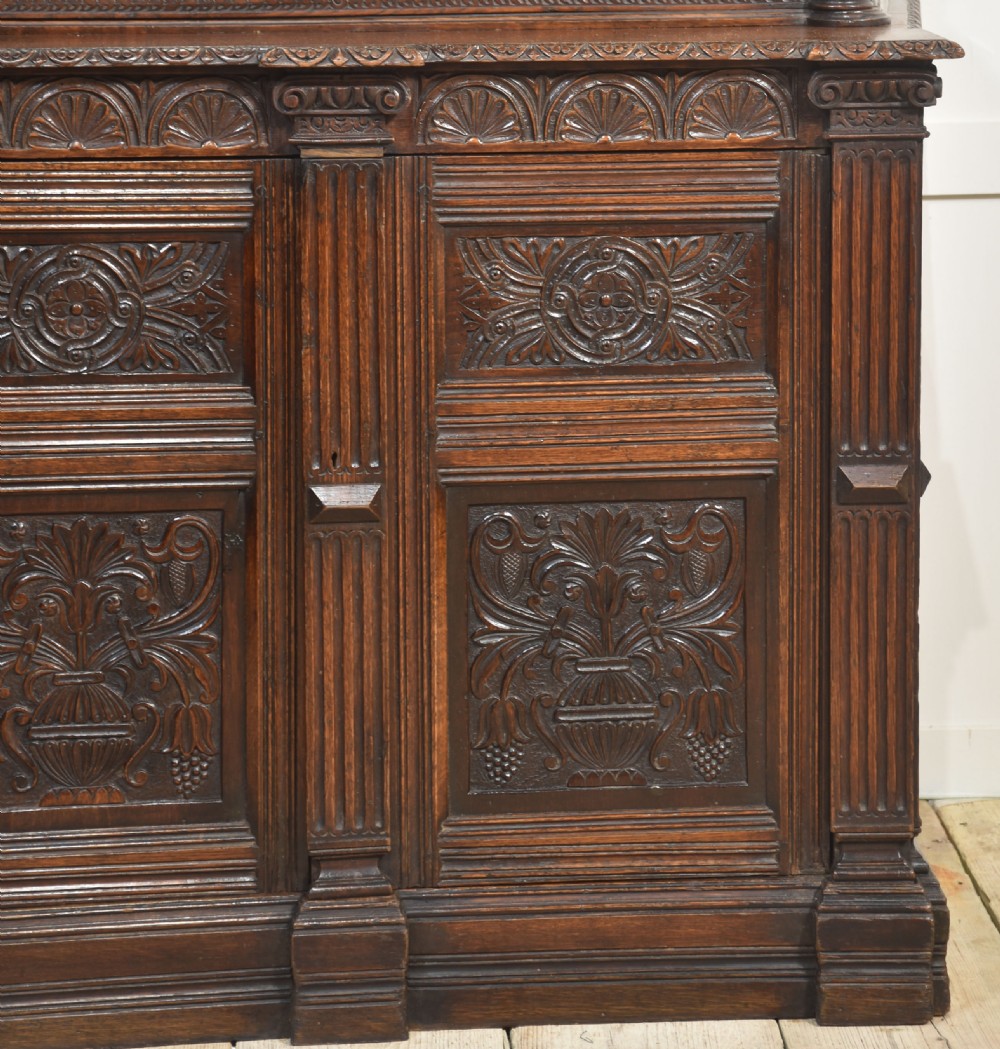 A dark carved oak display cabinet featuring a large upper glazed section with an arched spindle gallery top above two deeply relief-carved cupboard doors. View II
