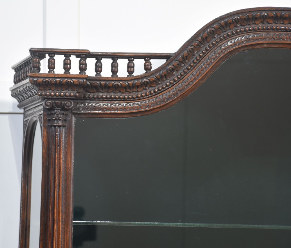 A dark carved oak display cabinet featuring a large upper glazed section with an arched spindle gallery top above two deeply relief-carved cupboard doors. View VIII