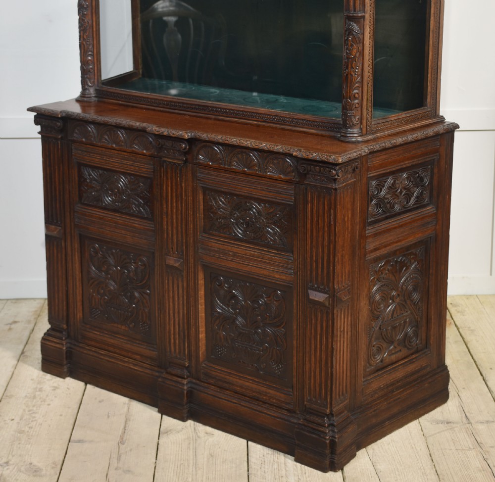 A dark carved oak display cabinet featuring a large upper glazed section with an arched spindle gallery top above two deeply relief-carved cupboard doors. View X
