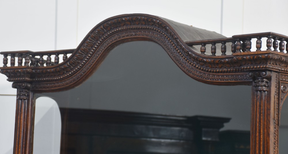 A dark carved oak display cabinet featuring a large upper glazed section with an arched spindle gallery top above two deeply relief-carved cupboard doors. View XII