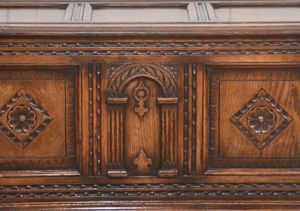 A dark carved oak coffer featuring three decorative front panels, turned legs, and a lower stretcher base. View III