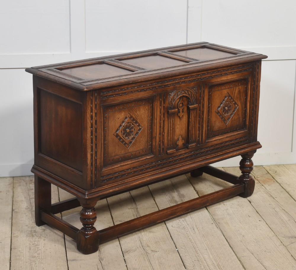 A dark carved oak coffer featuring three decorative front panels, turned legs, and a lower stretcher base. View V