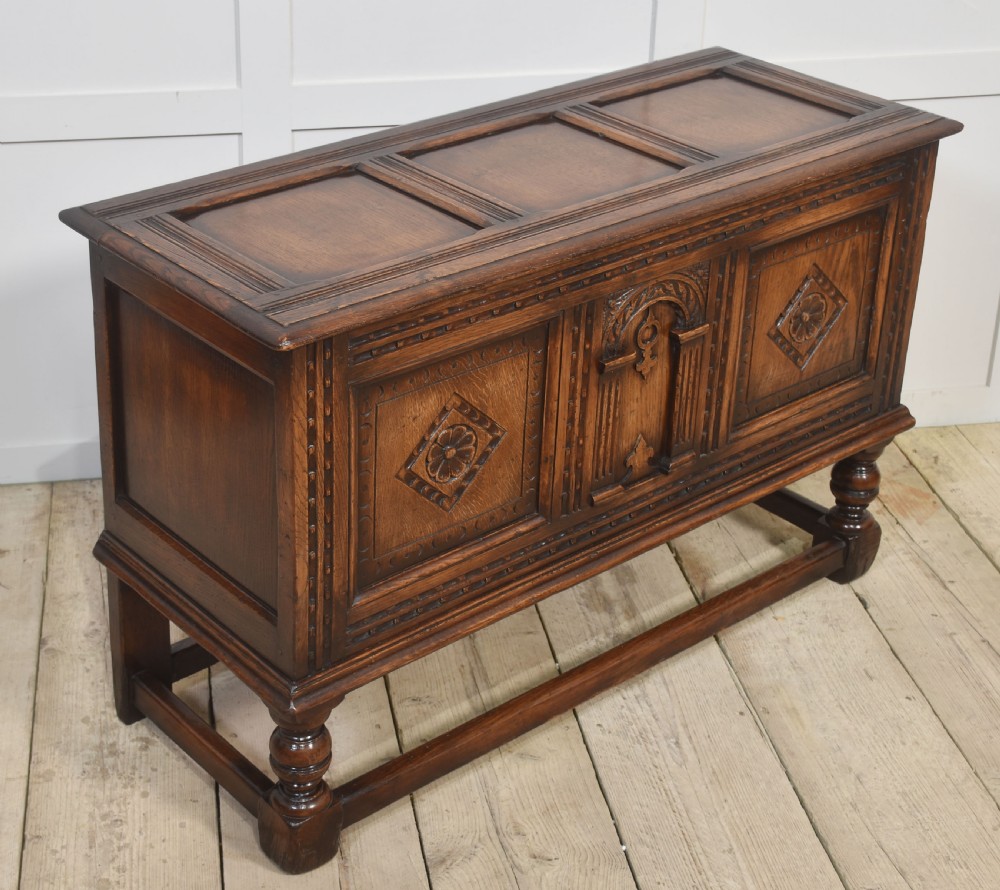 A dark carved oak coffer featuring three decorative front panels, turned legs, and a lower stretcher base. View VIII