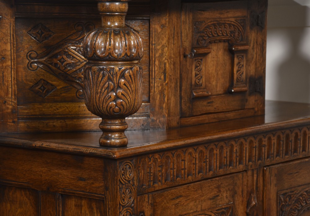 A two-tiered carved oak court cupboard featuring recessed upper doors flanked by bulbous turned supports and lower panelled doors adorned with carved arches. View XII