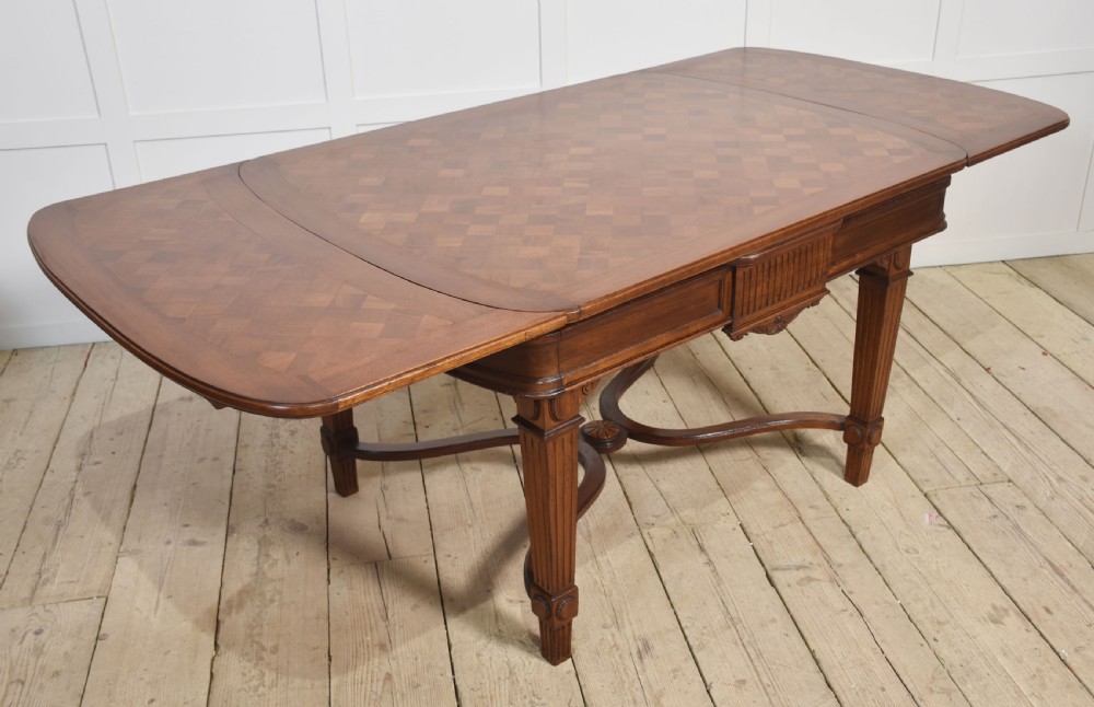 A walnut parquetry draw-leaf dining table featuring a geometric diamond-pattern top, fluted tapered legs, and a curved stretcher base.