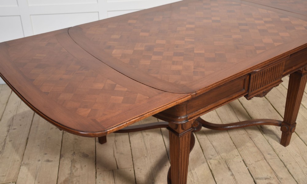 A walnut parquetry draw-leaf dining table featuring a geometric diamond-pattern top, fluted tapered legs, and a curved stretcher base. View VII