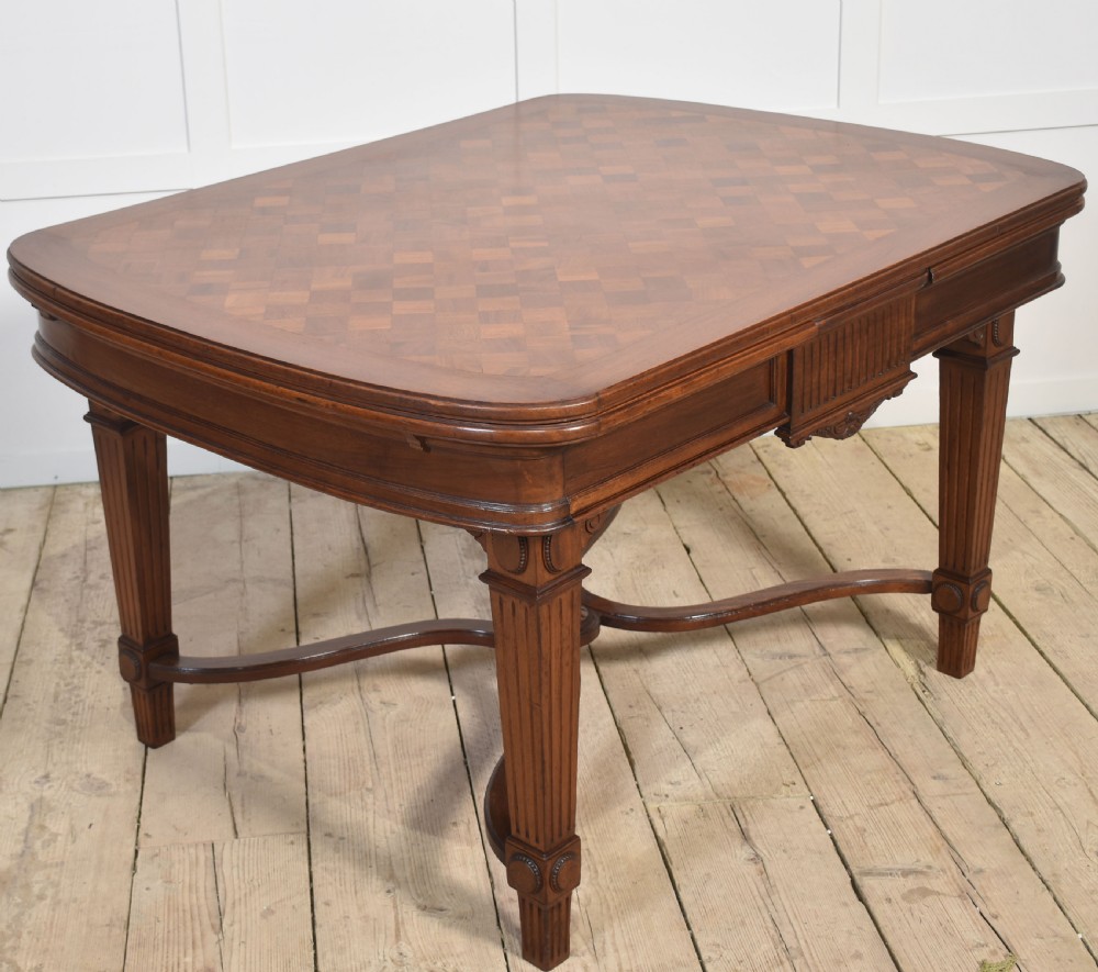 A walnut parquetry draw-leaf dining table featuring a geometric diamond-pattern top, fluted tapered legs, and a curved stretcher base. View XII