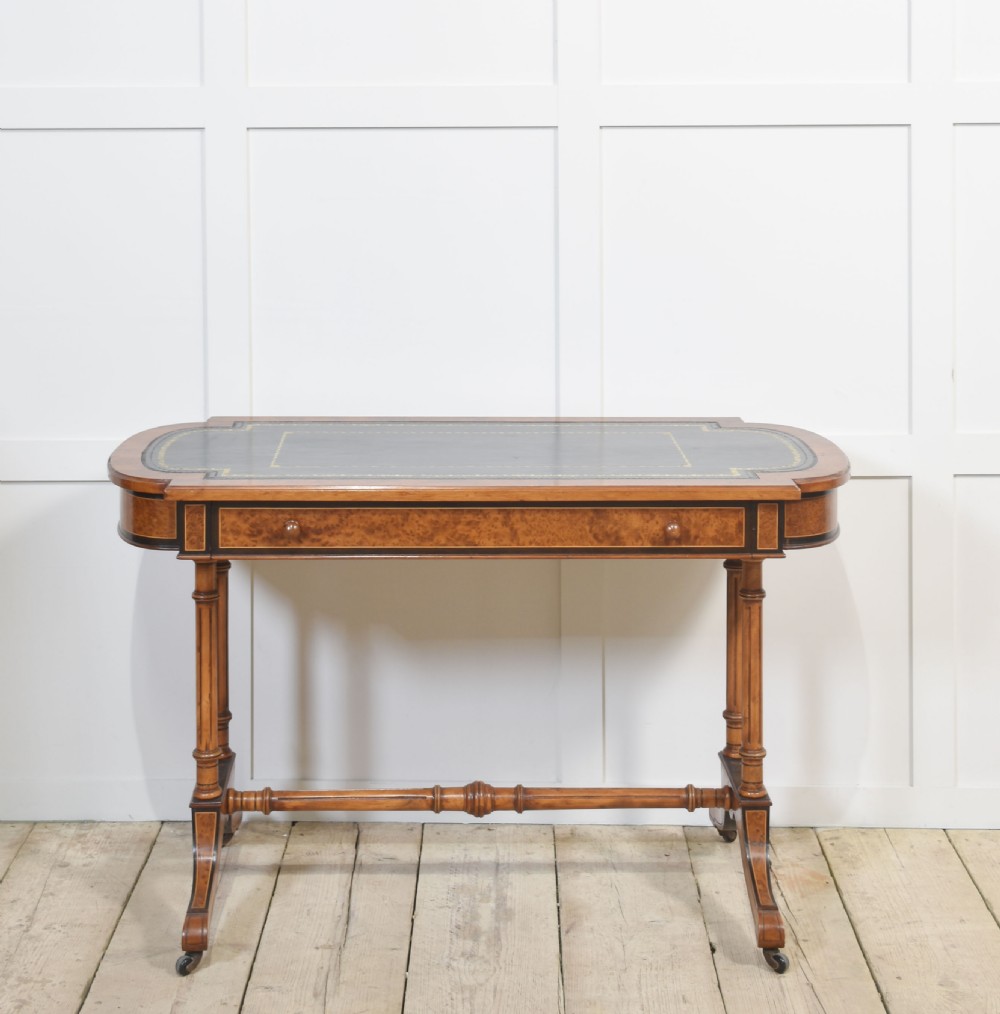 A Victorian burr walnut writing table featuring a green leather inset top with gold tooling, a single drawer, turned legs, a central stretcher, and brass castors.