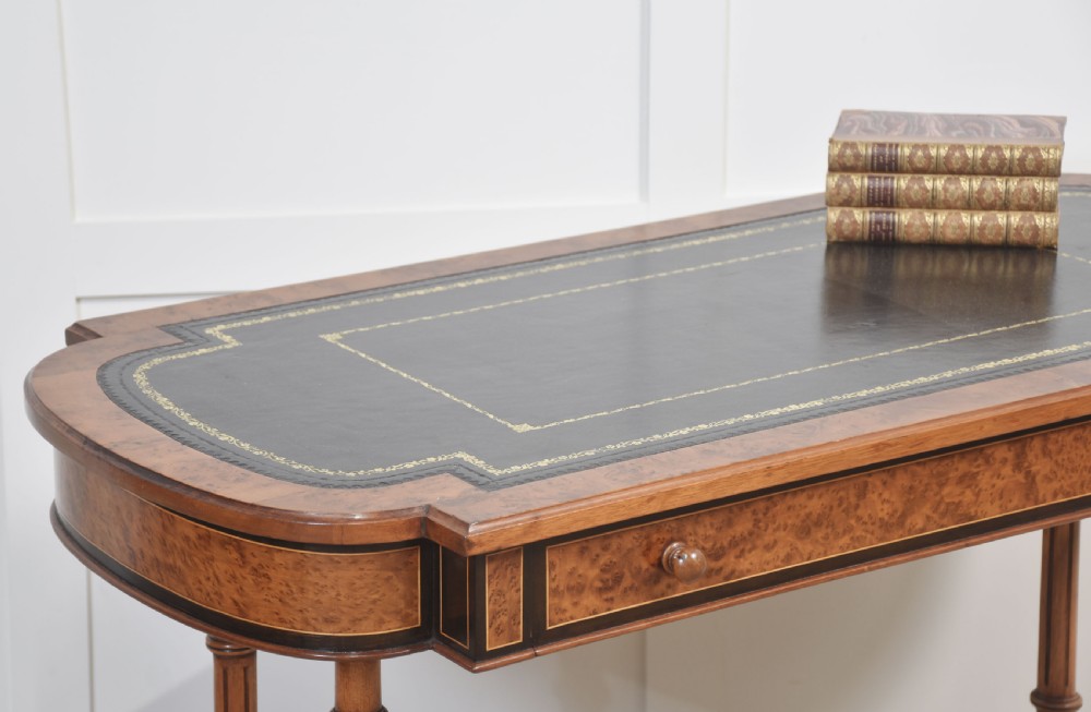 A Victorian burr walnut writing table featuring a green leather inset top with gold tooling, a single drawer, turned legs, a central stretcher, and brass castors. View V