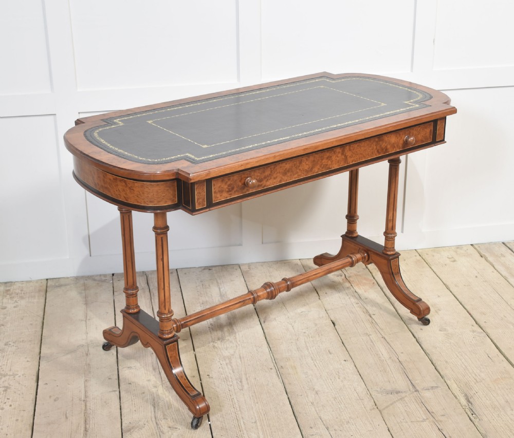 A Victorian burr walnut writing table featuring a green leather inset top with gold tooling, a single drawer, turned legs, a central stretcher, and brass castors. View VI