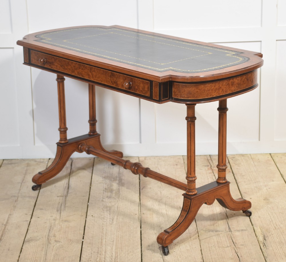 A Victorian burr walnut writing table featuring a green leather inset top with gold tooling, a single drawer, turned legs, a central stretcher, and brass castors. View X