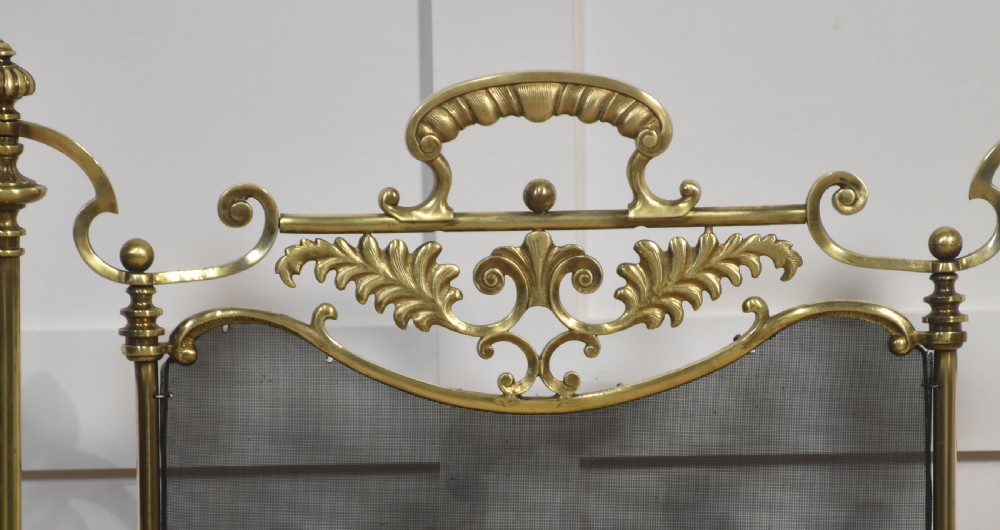 A brass fire screen featuring a wire mesh panel, decorative scrollwork framing, a top handle, and a central applied floral urn motif. View III