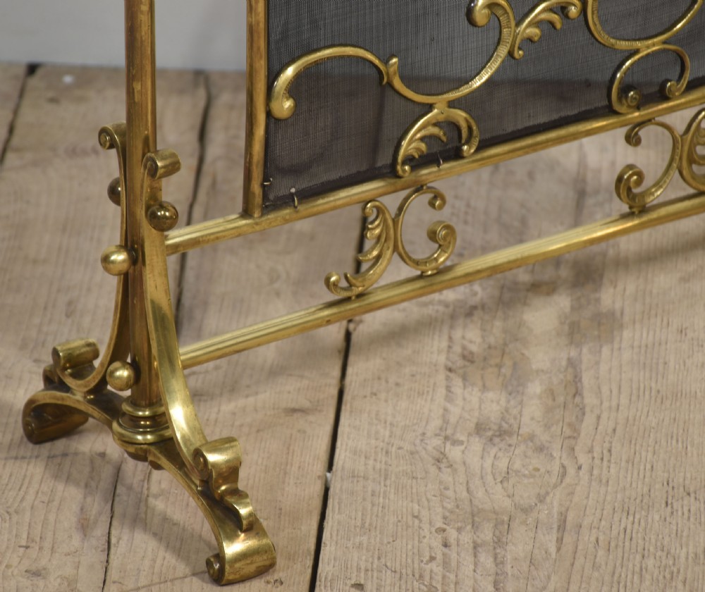 A brass fire screen featuring a wire mesh panel, decorative scrollwork framing, a top handle, and a central applied floral urn motif. View IV