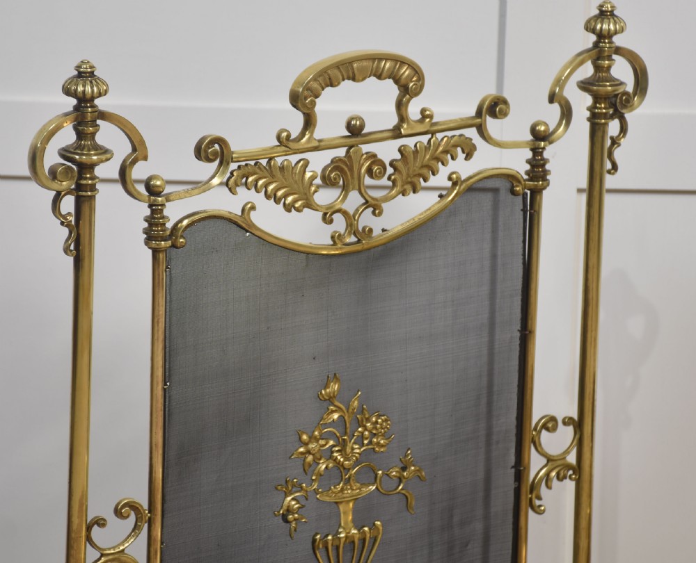 A brass fire screen featuring a wire mesh panel, decorative scrollwork framing, a top handle, and a central applied floral urn motif. View VI