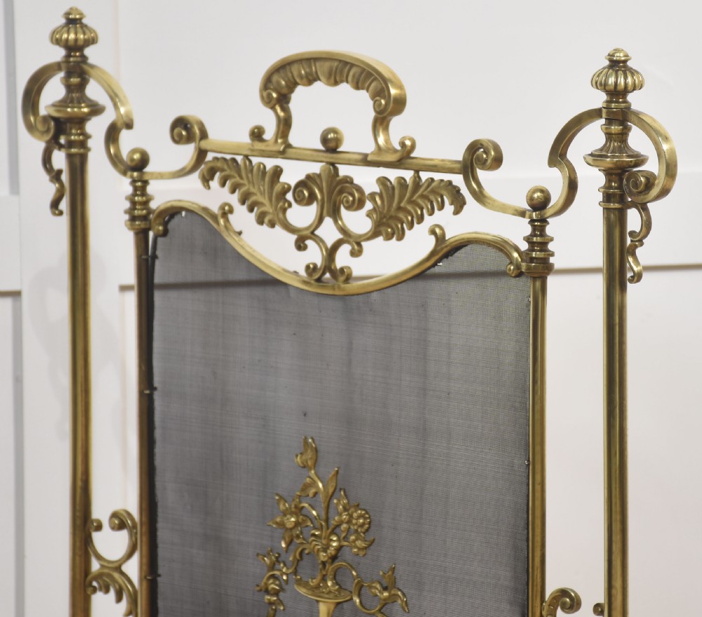 A brass fire screen featuring a wire mesh panel, decorative scrollwork framing, a top handle, and a central applied floral urn motif. View VIII