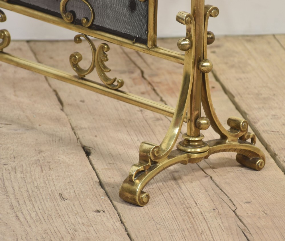 A brass fire screen featuring a wire mesh panel, decorative scrollwork framing, a top handle, and a central applied floral urn motif. View IX