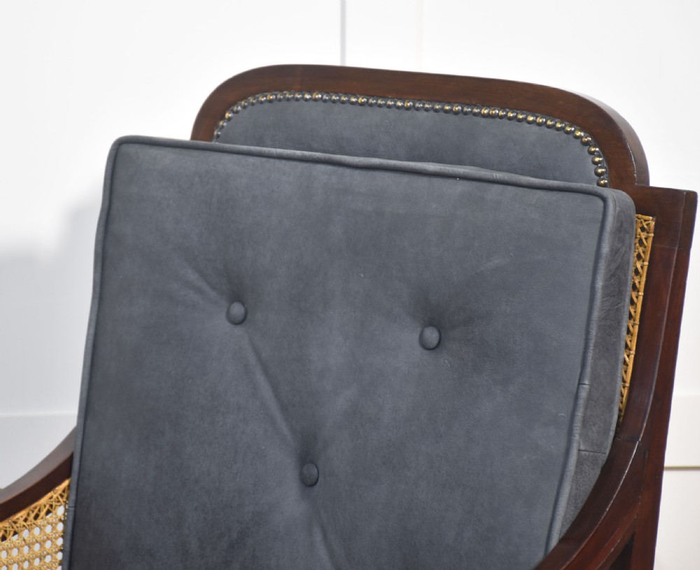 A Regency mahogany bergere armchair with hand-woven cane side panels, turned front legs on brass castors, and dark grey fabric cushions. View II