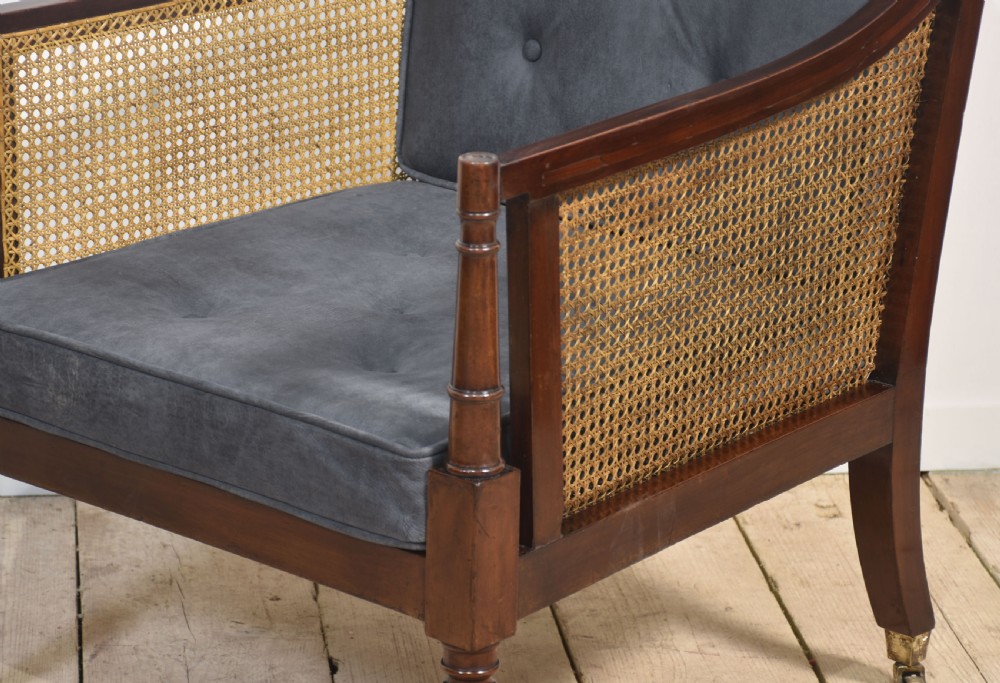 A Regency mahogany bergere armchair with hand-woven cane side panels, turned front legs on brass castors, and dark grey fabric cushions. View VI