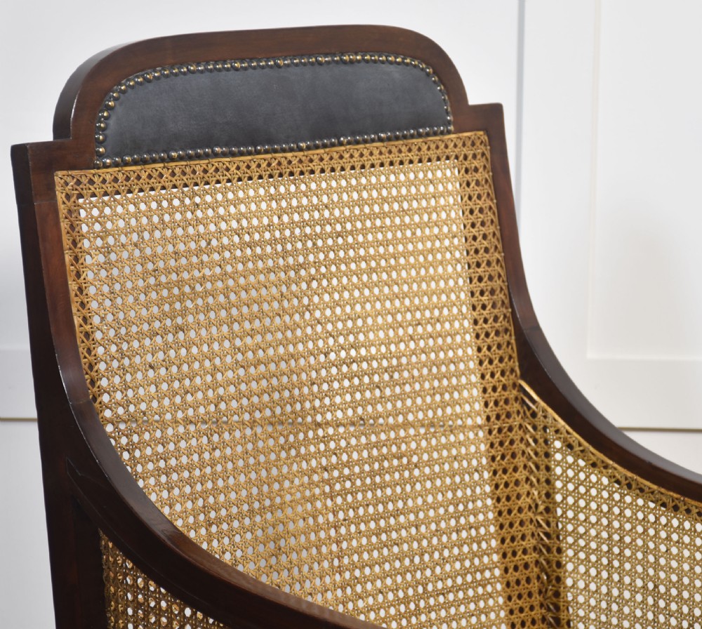 A Regency mahogany bergere armchair with hand-woven cane side panels, turned front legs on brass castors, and dark grey fabric cushions. View IX