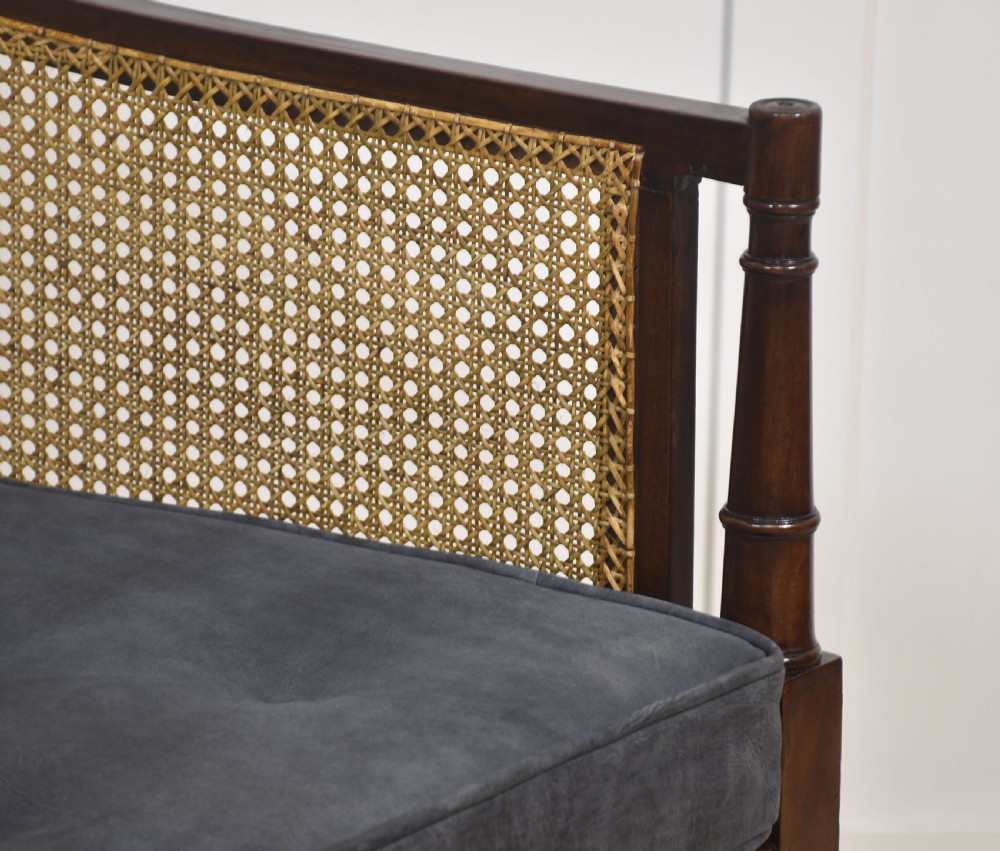 A Regency mahogany bergere armchair with hand-woven cane side panels, turned front legs on brass castors, and dark grey fabric cushions. View X