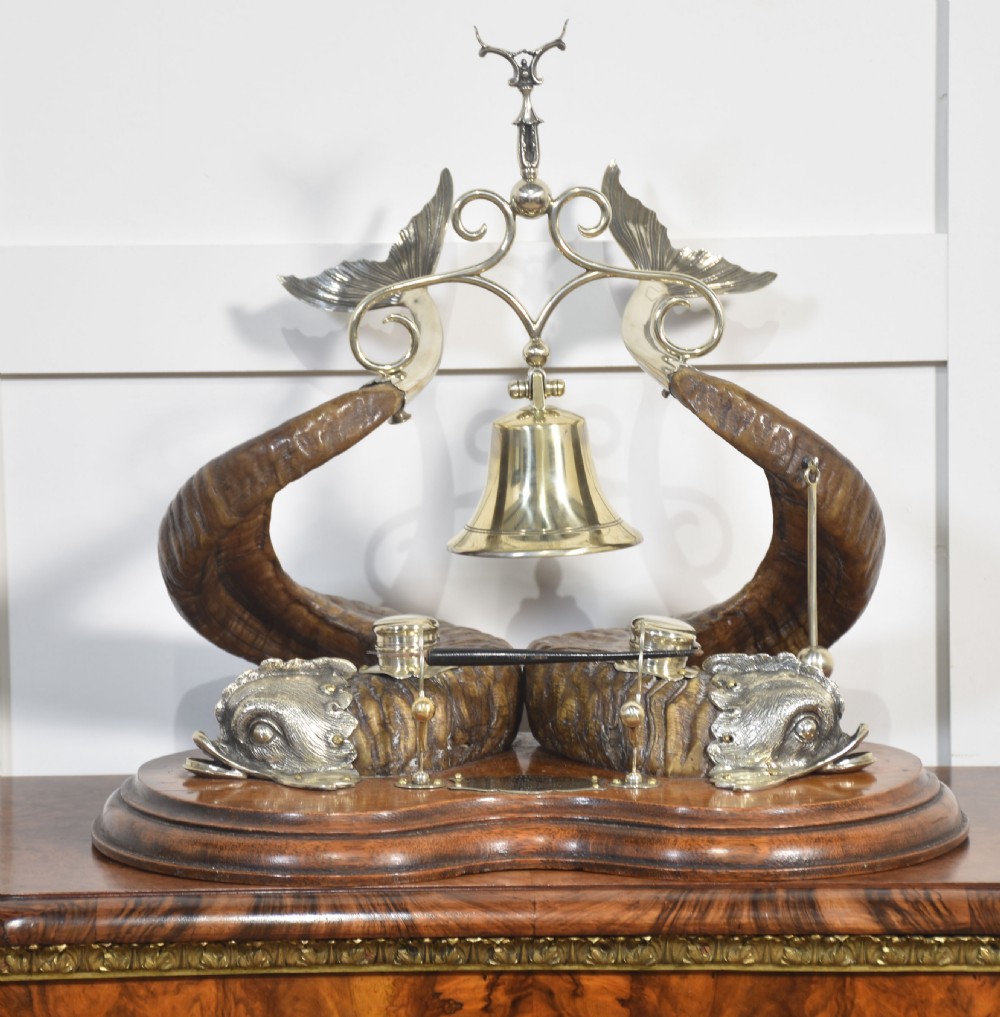 A 19th-century desk inkstand constructed from two curved natural horns, fitted with silvered dolphin-head mounts, inkwells, and a central suspended brass bell on a polished wooden base.