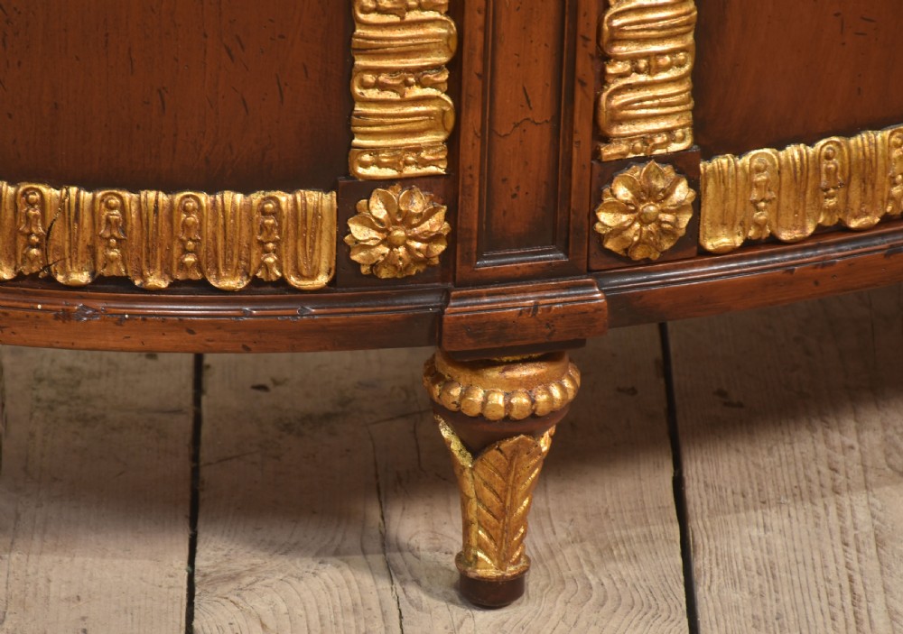 An Italian Neoclassical mahogany bowfront cabinet featuring a black marble top, marquetry frieze with foliate motifs, and carved parcel gilt trims. View X