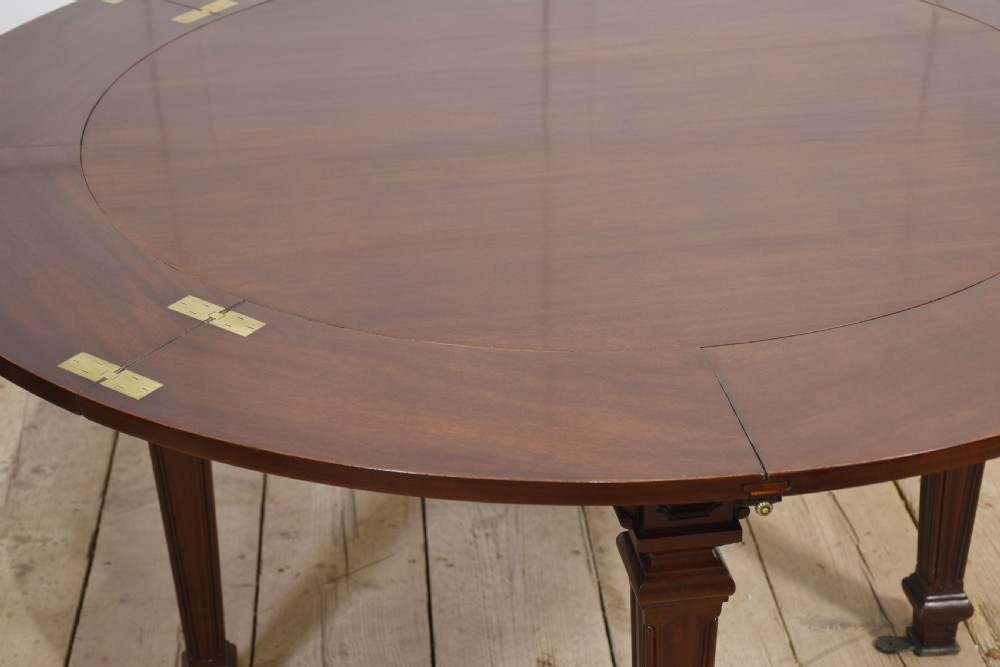 A dark mahogany circular dining table featuring outer leaf extension segments connected with brass hinges and supported by four fluted legs. View VI