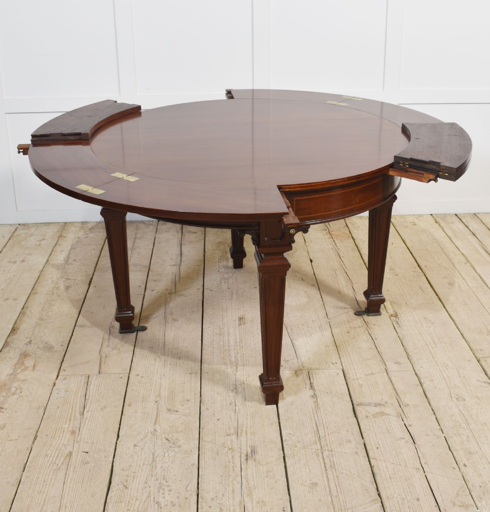 A dark mahogany circular dining table featuring outer leaf extension segments connected with brass hinges and supported by four fluted legs. View IX