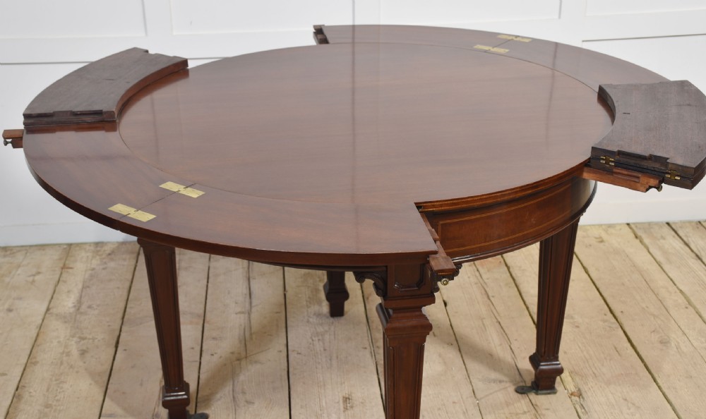 A dark mahogany circular dining table featuring outer leaf extension segments connected with brass hinges and supported by four fluted legs. View X