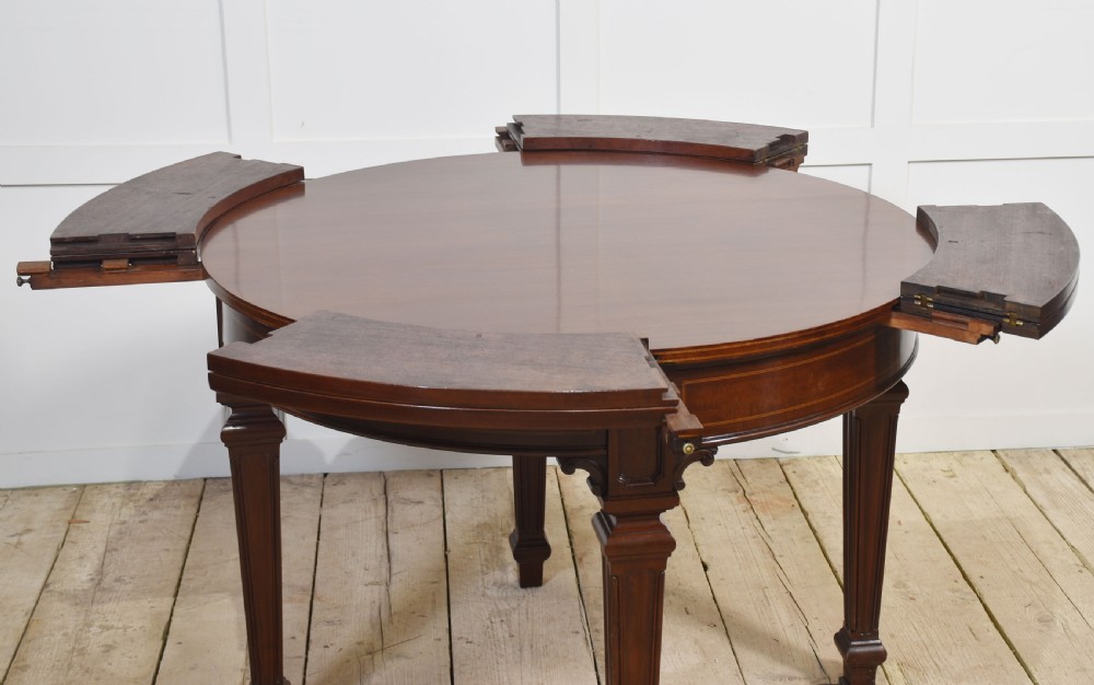 A dark mahogany circular dining table featuring outer leaf extension segments connected with brass hinges and supported by four fluted legs. View XI